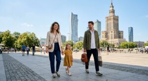 A high-quality editorial photograph of a stylish young family walking through a sunny Warsaw square,